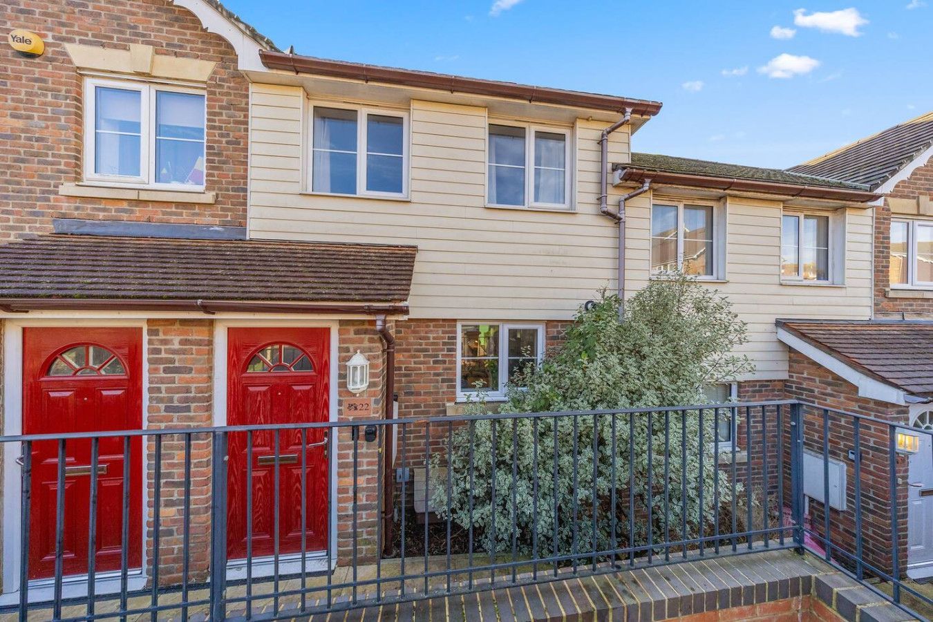 A row of townhouses with red doors, brick and siding exterior, and a small metal fence in the foreground under a blue sky.
