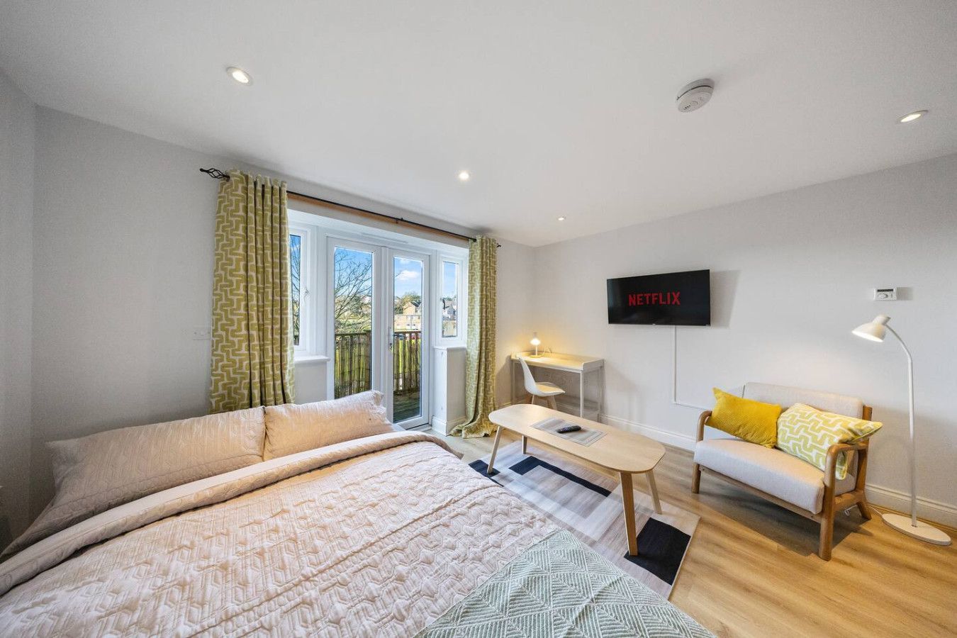 A bright bedroom featuring a bed, small sofa, coffee table, desk, and French doors with yellow patterned curtains.
