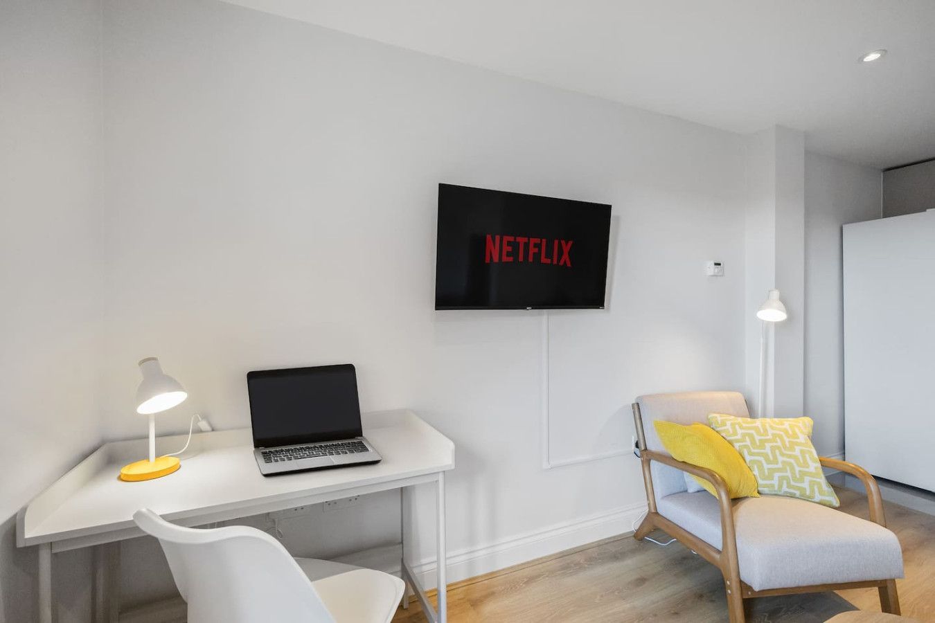 A modern room with a white desk, laptop, lamp, Netflix on a wall-mounted TV, and a chair with yellow and patterned pillows.