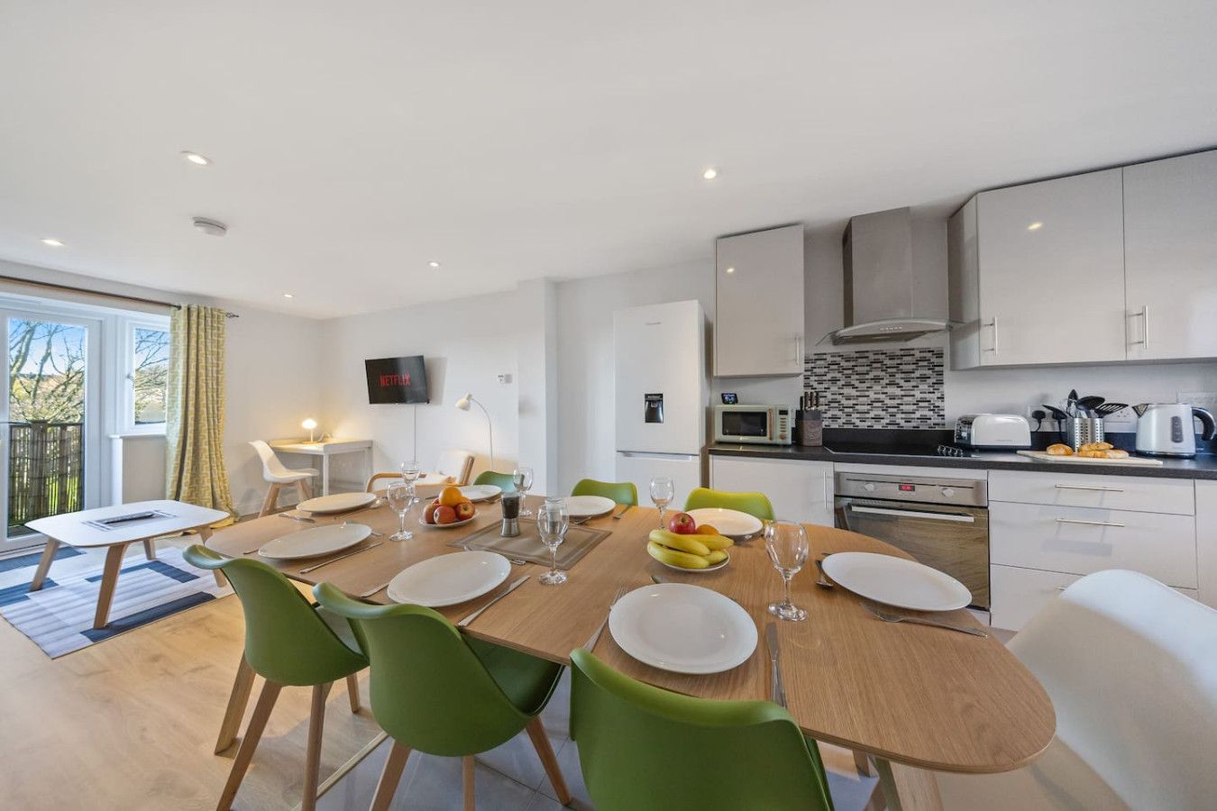A bright, modern kitchen and dining area featuring a light wood table with six green chairs and a balcony view.