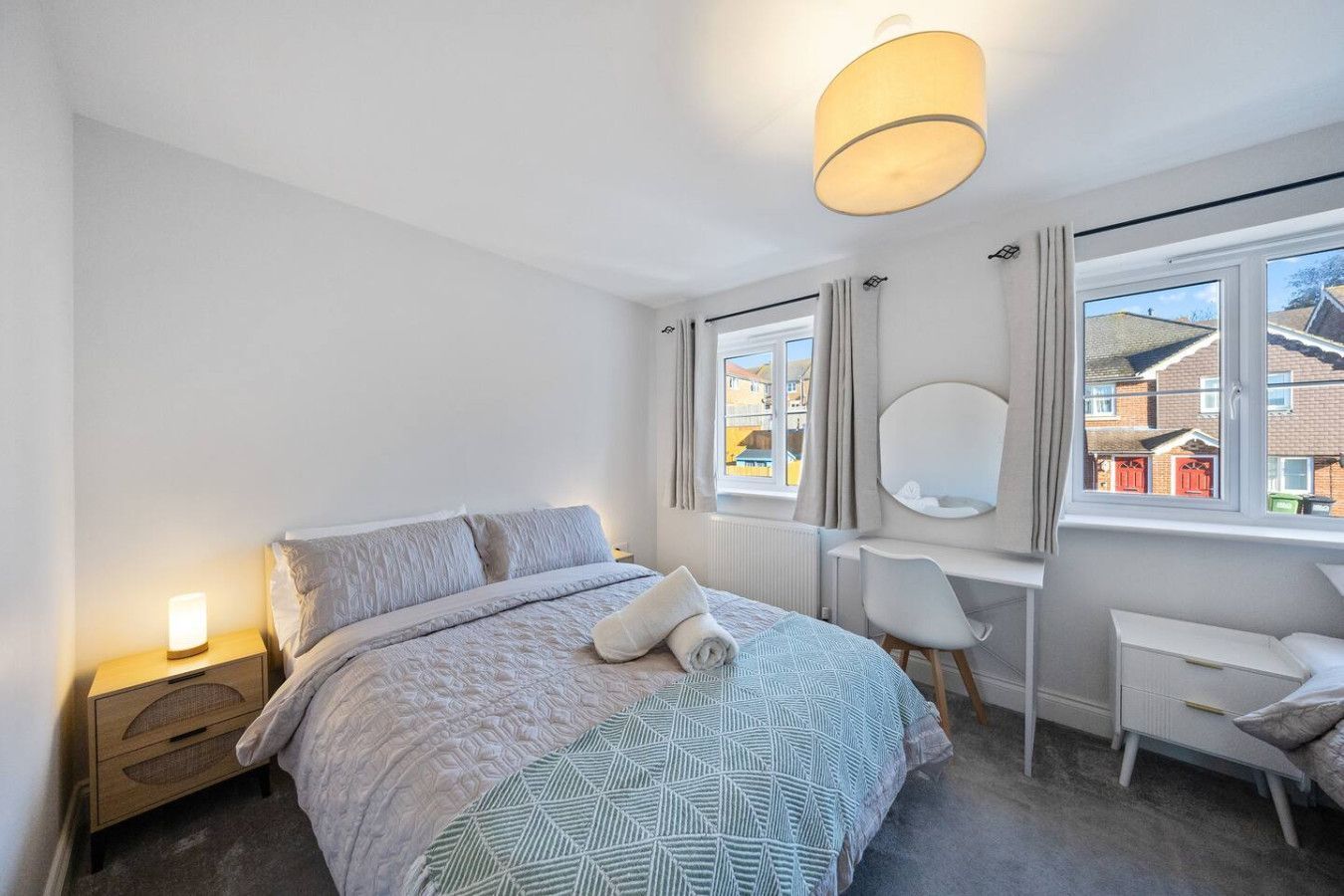 A bright bedroom with a queen bed featuring a patterned gray quilt, a bedside lamp, a vanity desk, and a window.