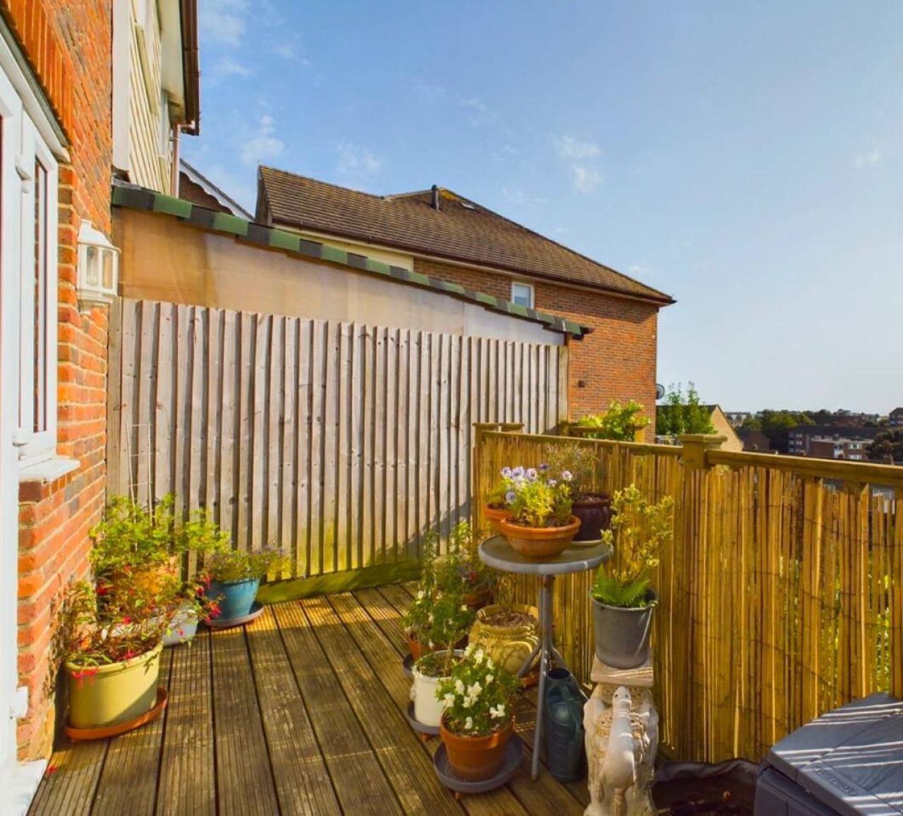 A wooden deck balcony features potted plants, a small bistro table, and a bamboo privacy screen under a clear blue sky.