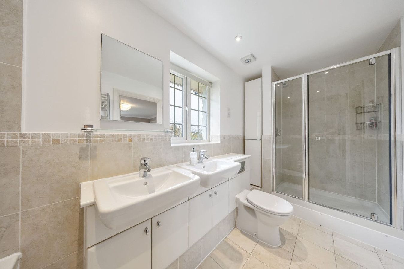 Modern bathroom with a white vanity sink, a large mirror, a toilet, and a glass-enclosed shower against beige tile walls.