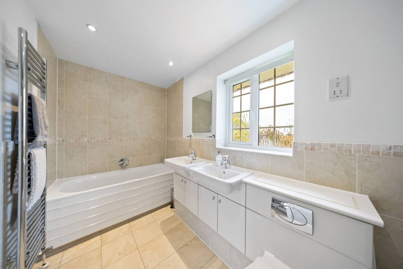 A bright bathroom with a white bathtub, tiled walls, double vanity, chrome towel radiator, and a window.