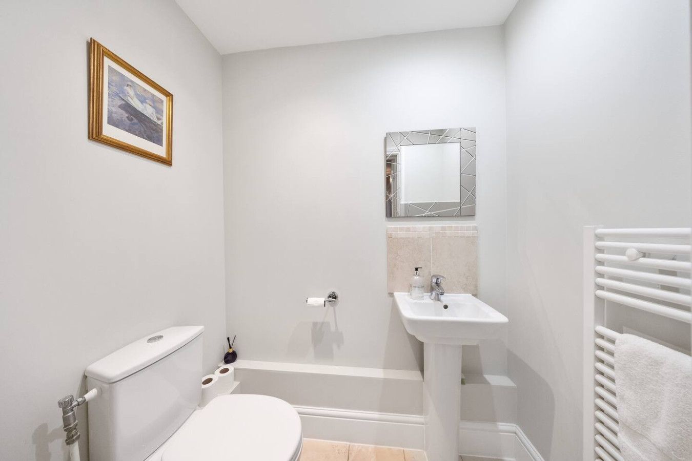 A minimalist white powder room featuring a toilet, a pedestal sink with a small mirror above it, and a towel radiator.