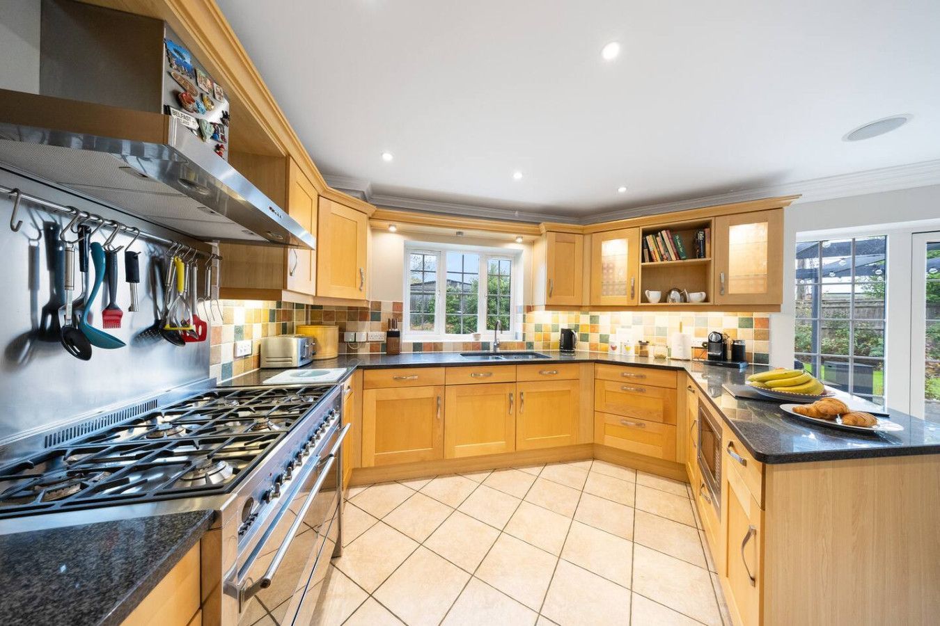 A spacious, well-lit kitchen featuring light-wood cabinetry, a large gas range, tiled floors, and a corner window.