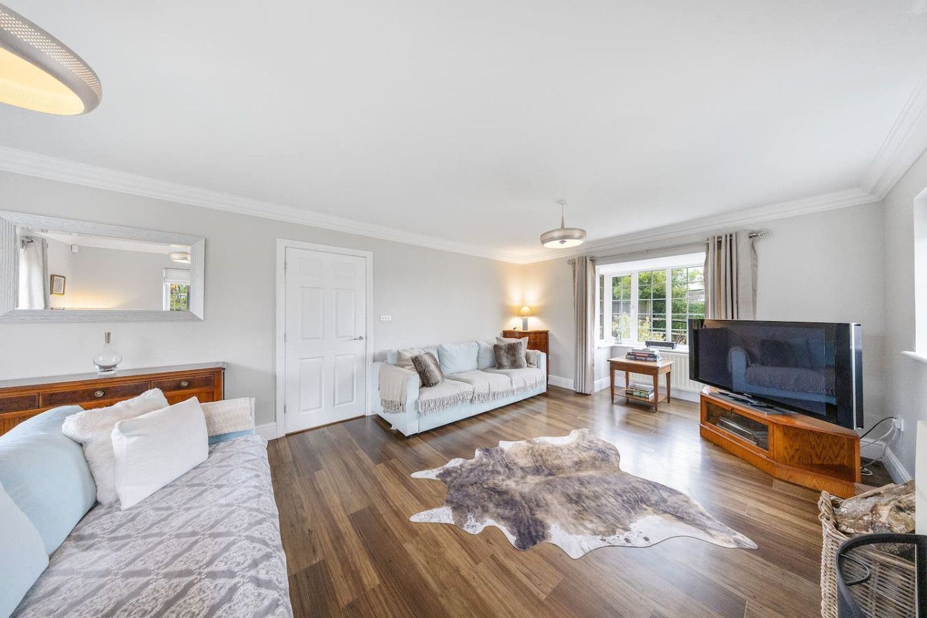 A bright, modern living room with light-colored sofas, a patterned rug, wooden floors, and a television on a wood stand.