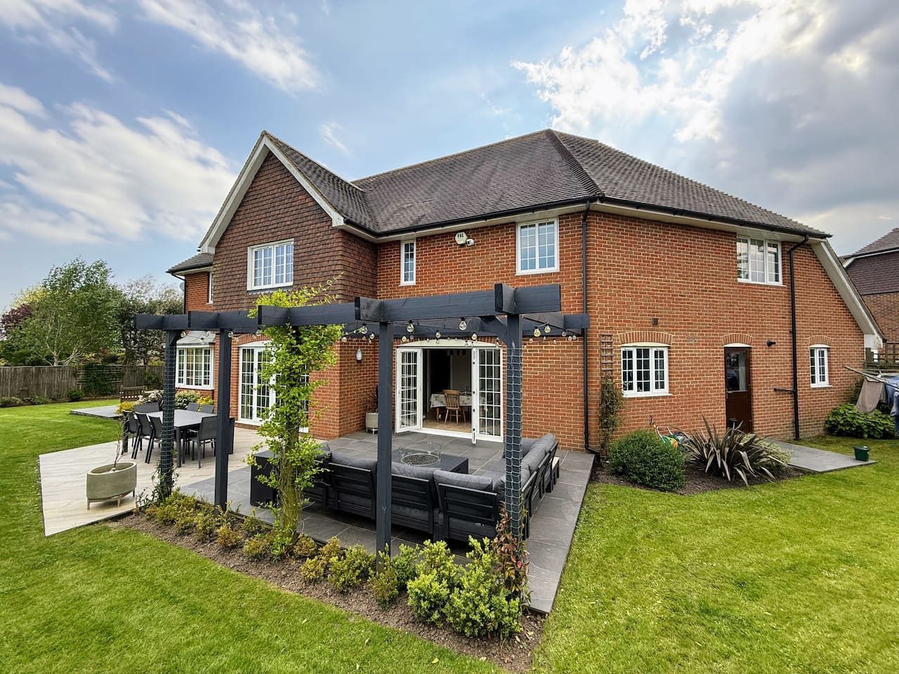 A backyard view of a red-brick, two-story house featuring a patio with a dark pergola, outdoor furniture, and a lawn.