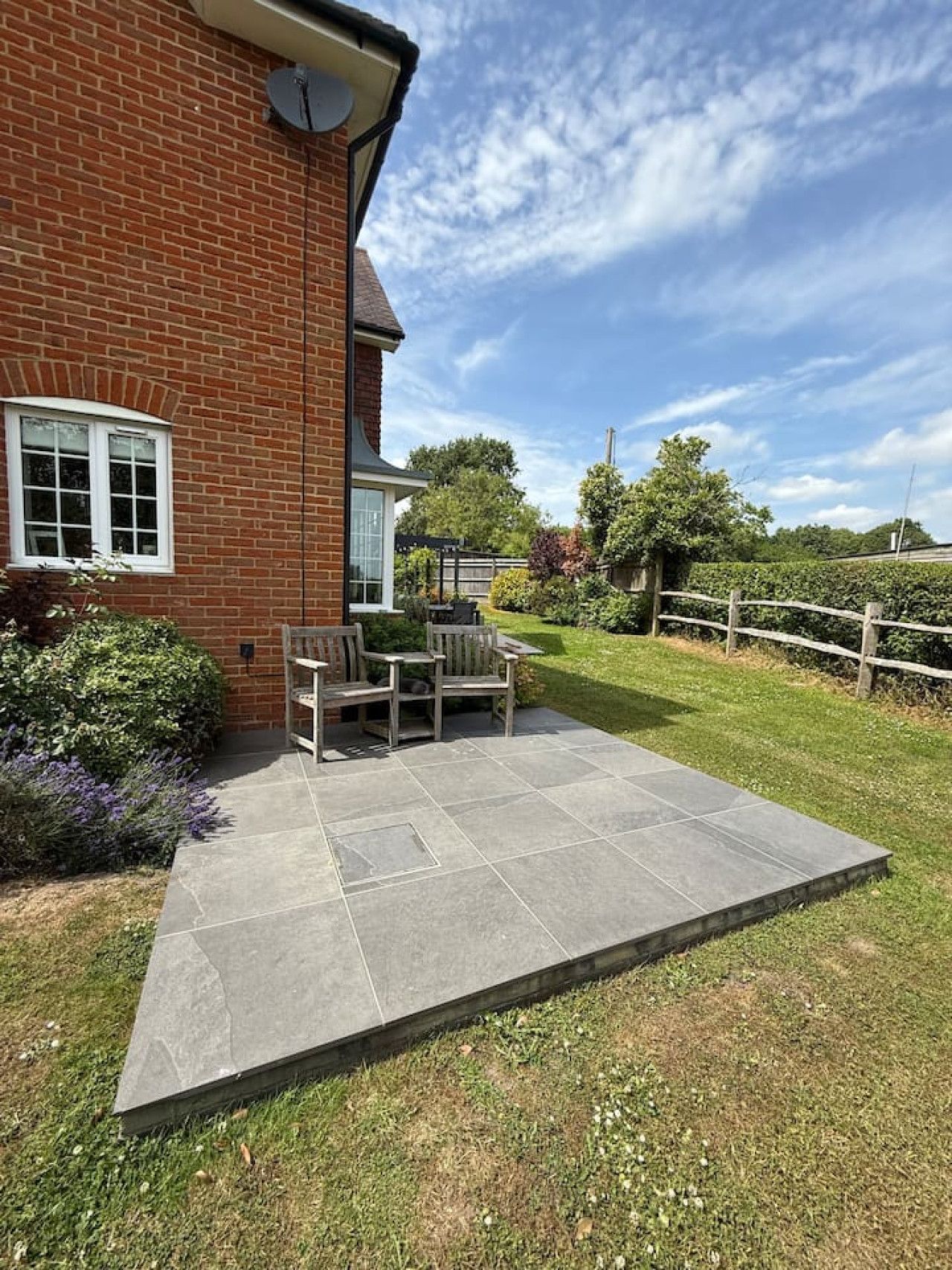 A grey stone patio with two chairs and a table sits next to a brick house in a green, sunny garden.