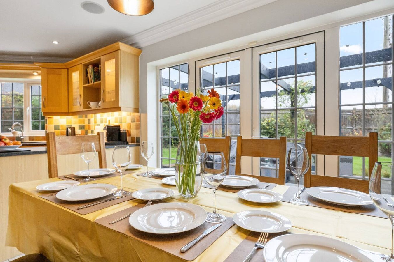 A formal dining table set with white plates, glasses, and a vibrant flower bouquet in a bright, wood-toned kitchen.