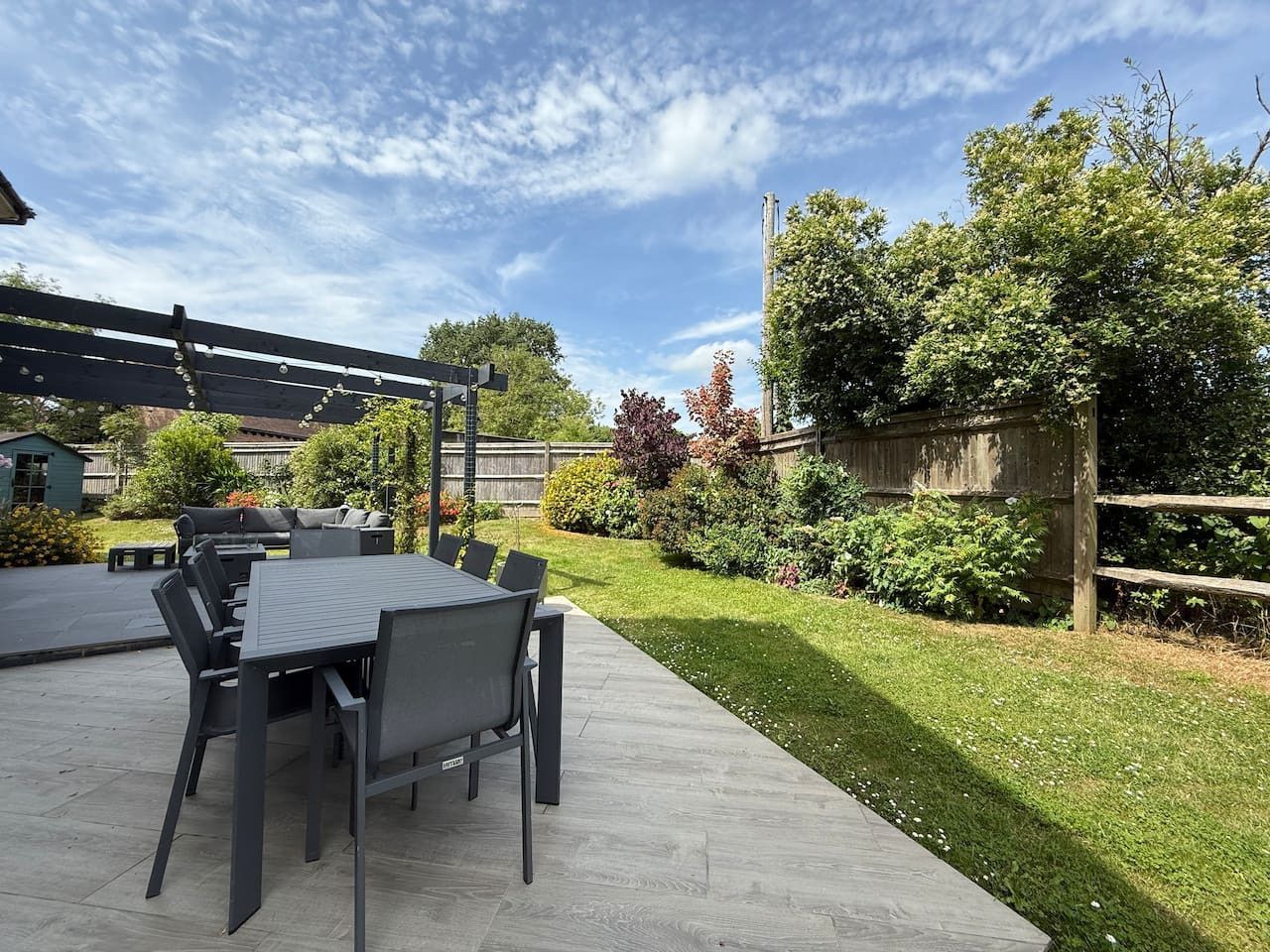A dark gray outdoor dining set with a pergola on a stone patio overlooking a green lawn and garden.