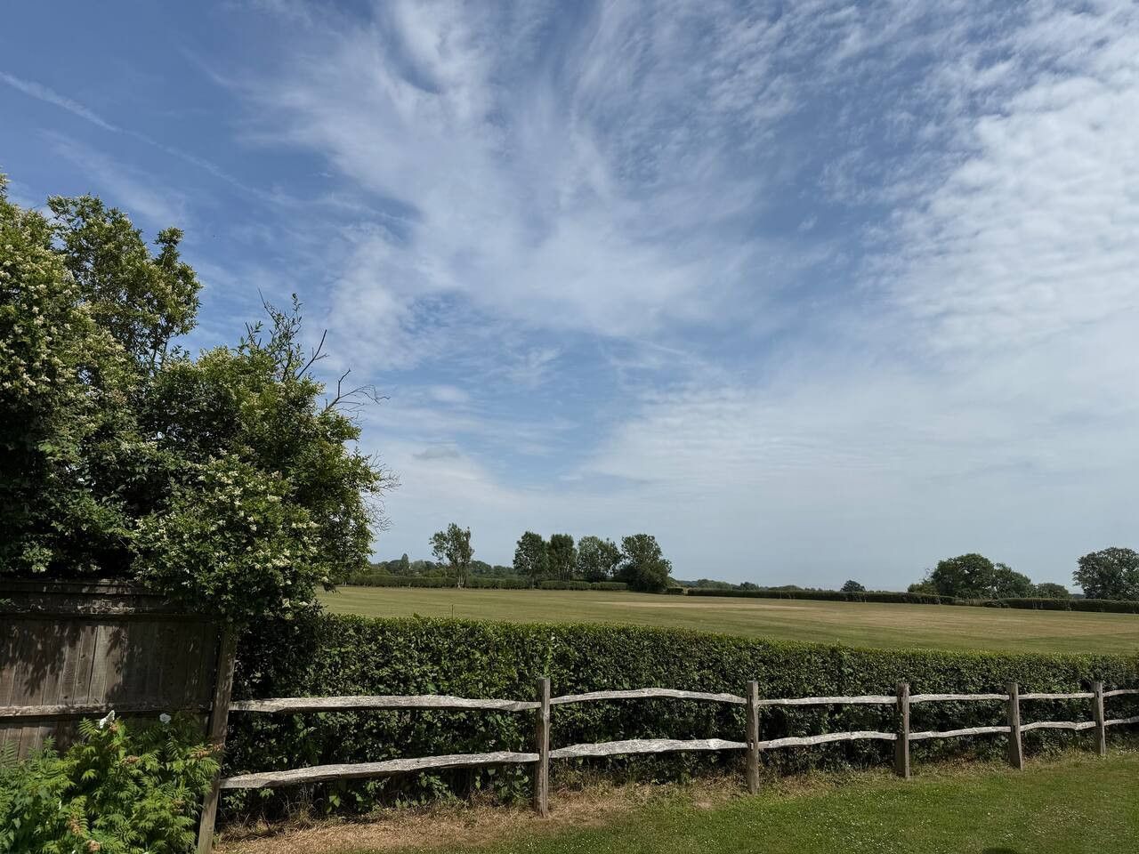 A rustic wooden fence borders a green meadow under a bright blue sky with wispy clouds.