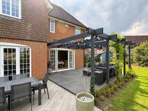 A brick house features a black pergola over a dark patio, with a dining set on light wooden decking and a green lawn.