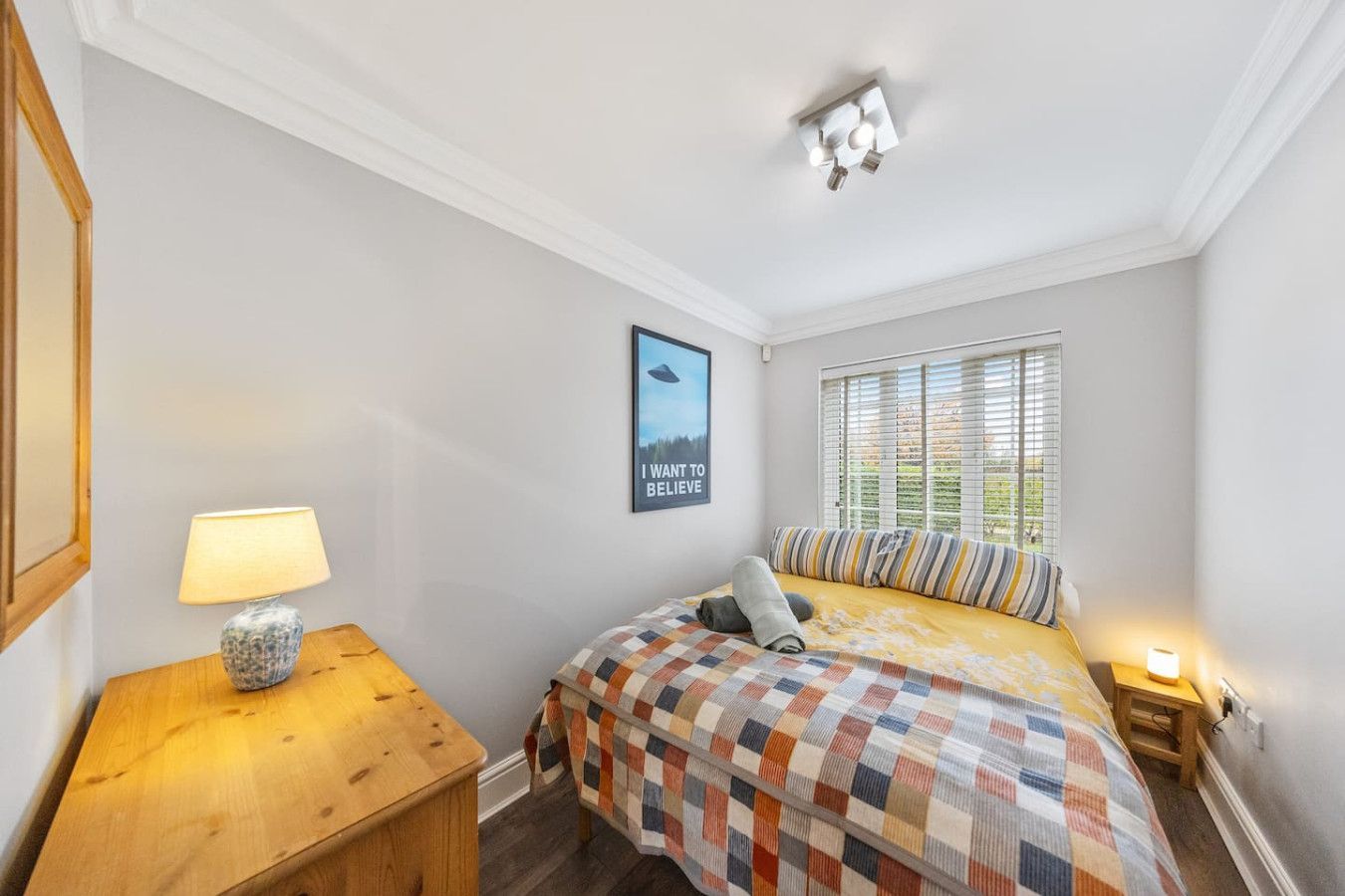 A small bedroom with a bed featuring a checkered quilt, a wooden dresser with a lamp, and a framed wall art piece.