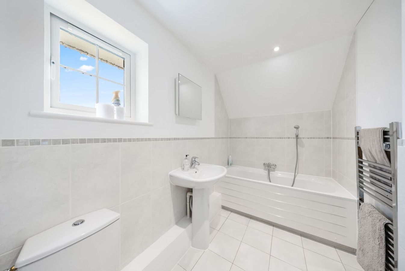 A modern, bright bathroom with white tile walls and floor, featuring a pedestal sink, bathtub, and a window.