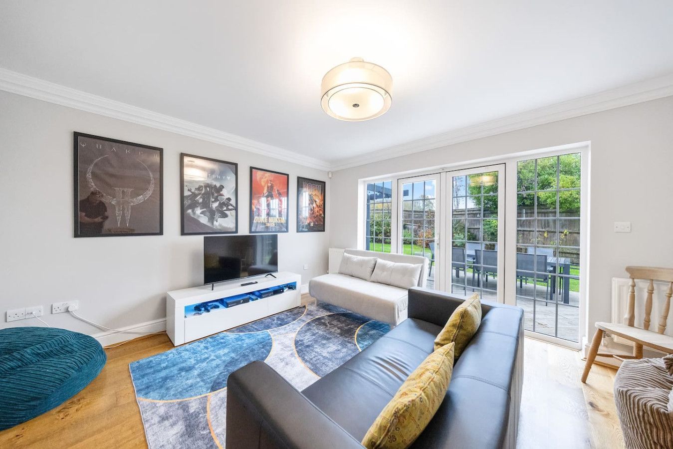 A brightly lit living room with a black leather sofa, white media unit, blue abstract rug, and glass patio doors.