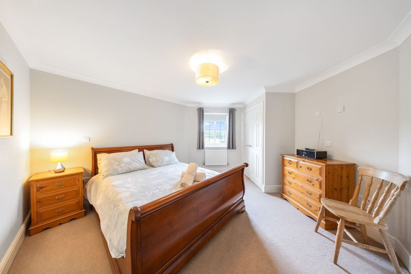 A bedroom with a wooden bed frame, bedside table, dresser, chair, and a window centered on the light-colored wall.