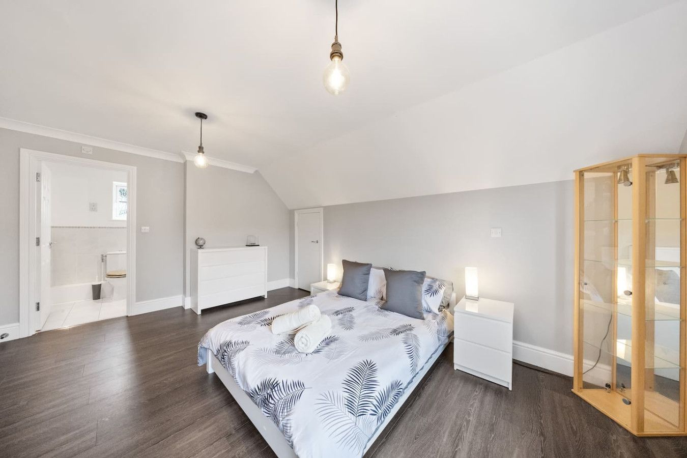 A bedroom with a queen bed, dark wood floors, white walls, a white dresser, and an open doorway to an ensuite bathroom.