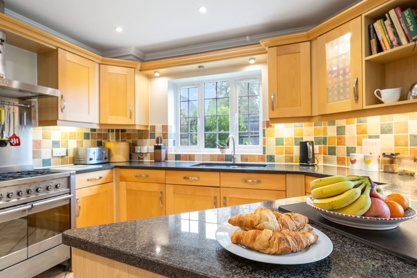 A bright kitchen with light wood cabinets, granite countertops, a tiled backsplash, and a bowl of fruit with croissants.