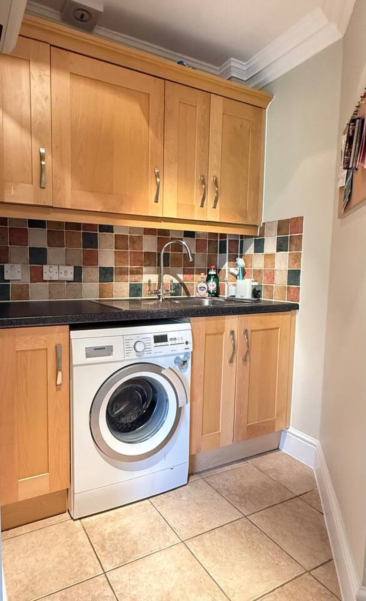 A small utility kitchen featuring a white washing machine, light wood cabinets, and a mosaic tile backsplash.