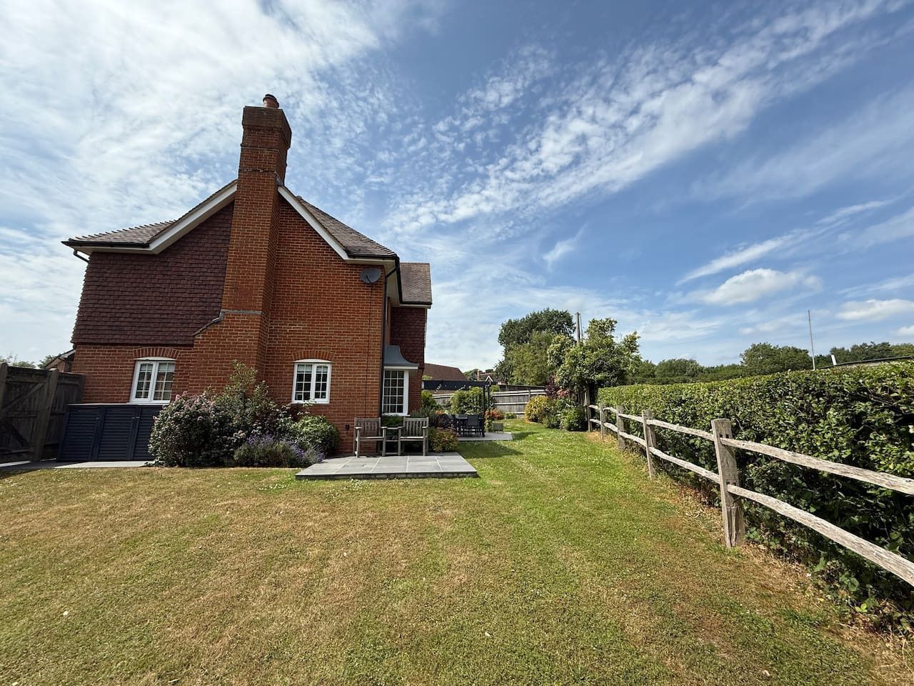 A red brick house with a tall chimney sits beside a grass lawn and a wooden split-rail fence under a bright blue sky.