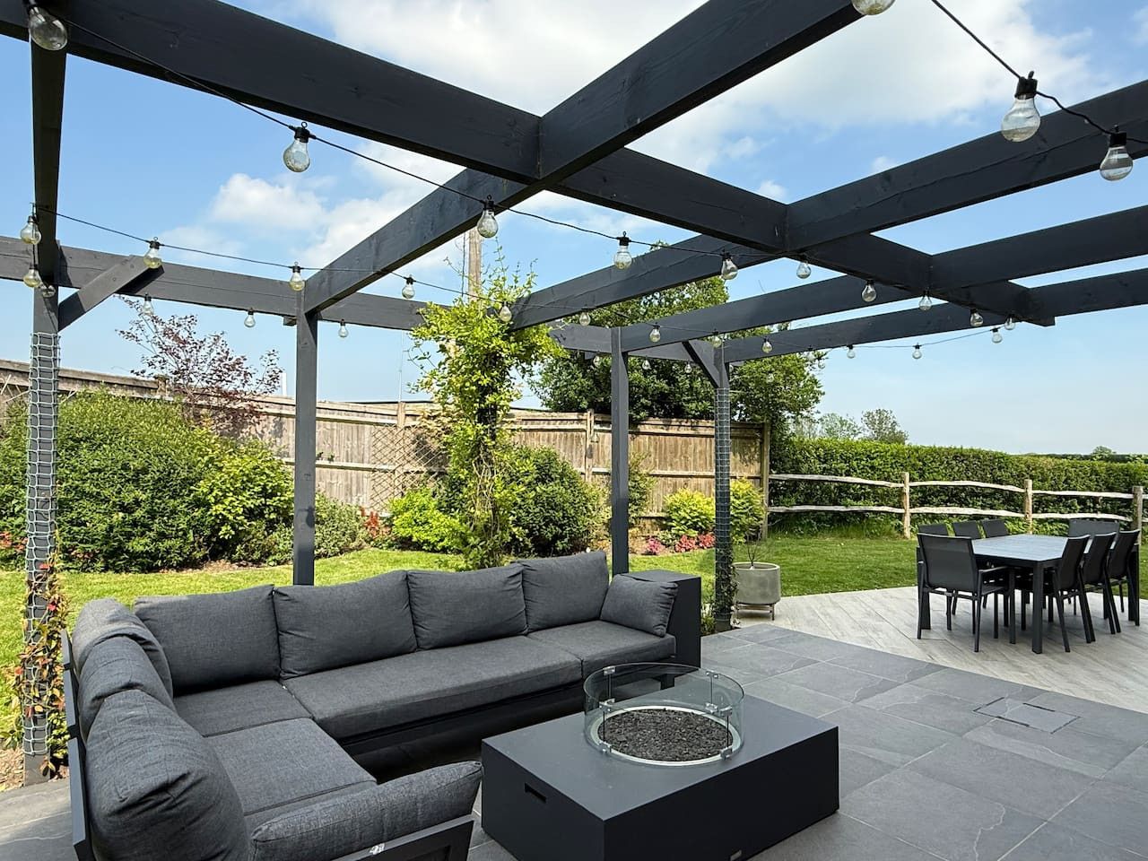 A modern patio with a dark grey pergola, comfortable sofa, fire pit table, and a dining set on a sunny day.