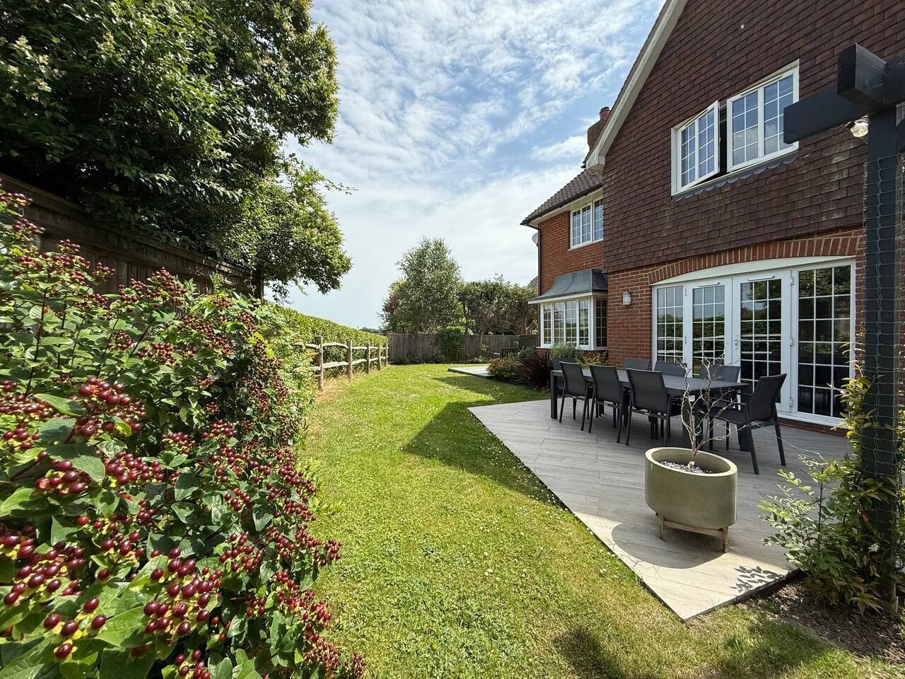 A brick house with French doors opens onto a stone patio and lawn, bordered by a large, berry-filled bush.