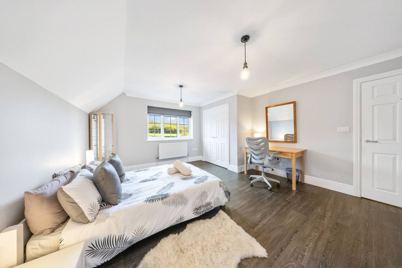 A bright bedroom with grey walls, a bed featuring a patterned duvet, a wooden desk with a chair, and a white door.