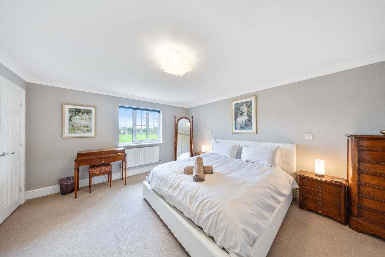 A well-lit, neutral-toned bedroom featuring a large white bed, wooden furniture, and a window overlooking greenery.