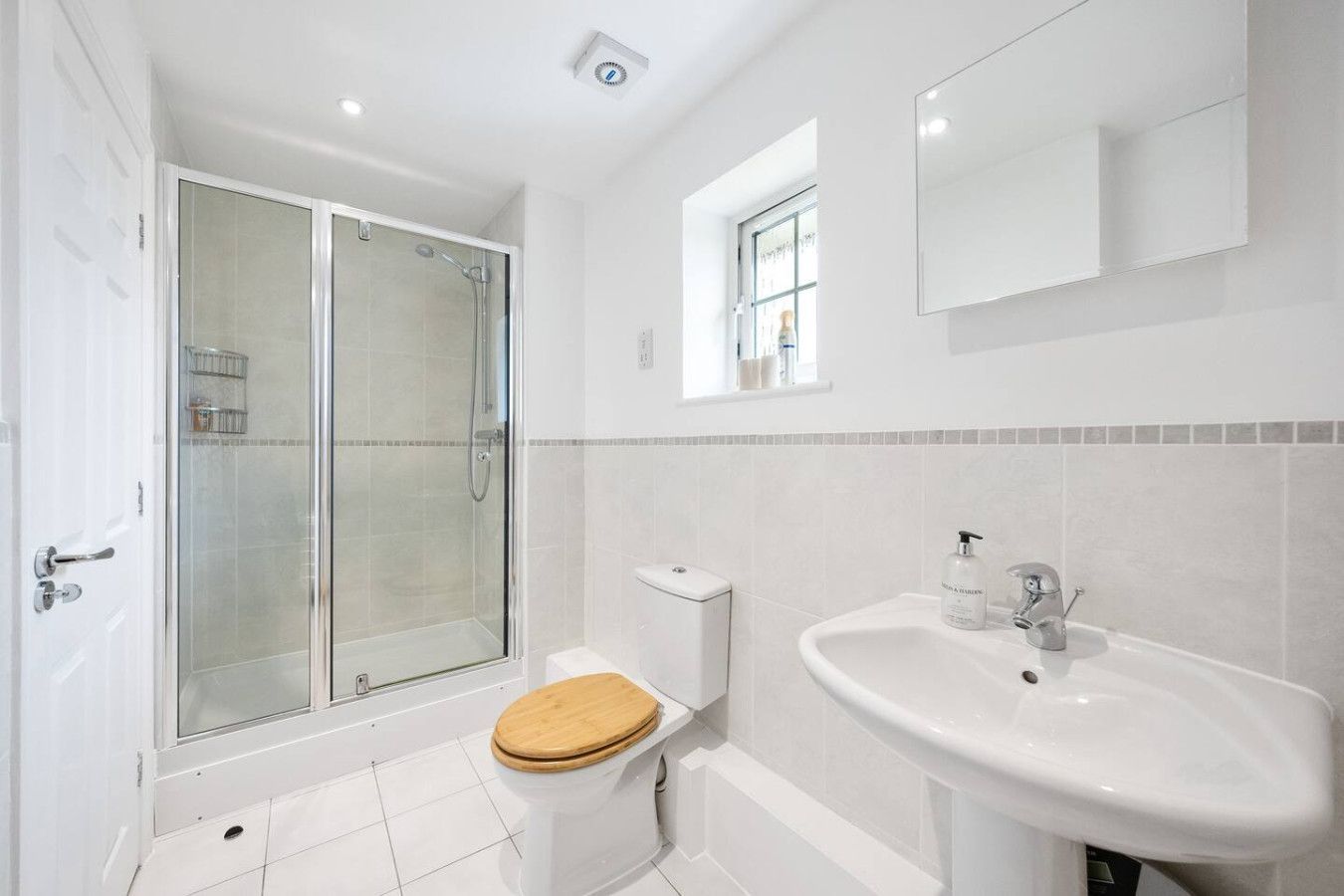 A brightly lit bathroom with a glass-enclosed shower, a white pedestal sink, a toilet with a wood seat, and white tiles.