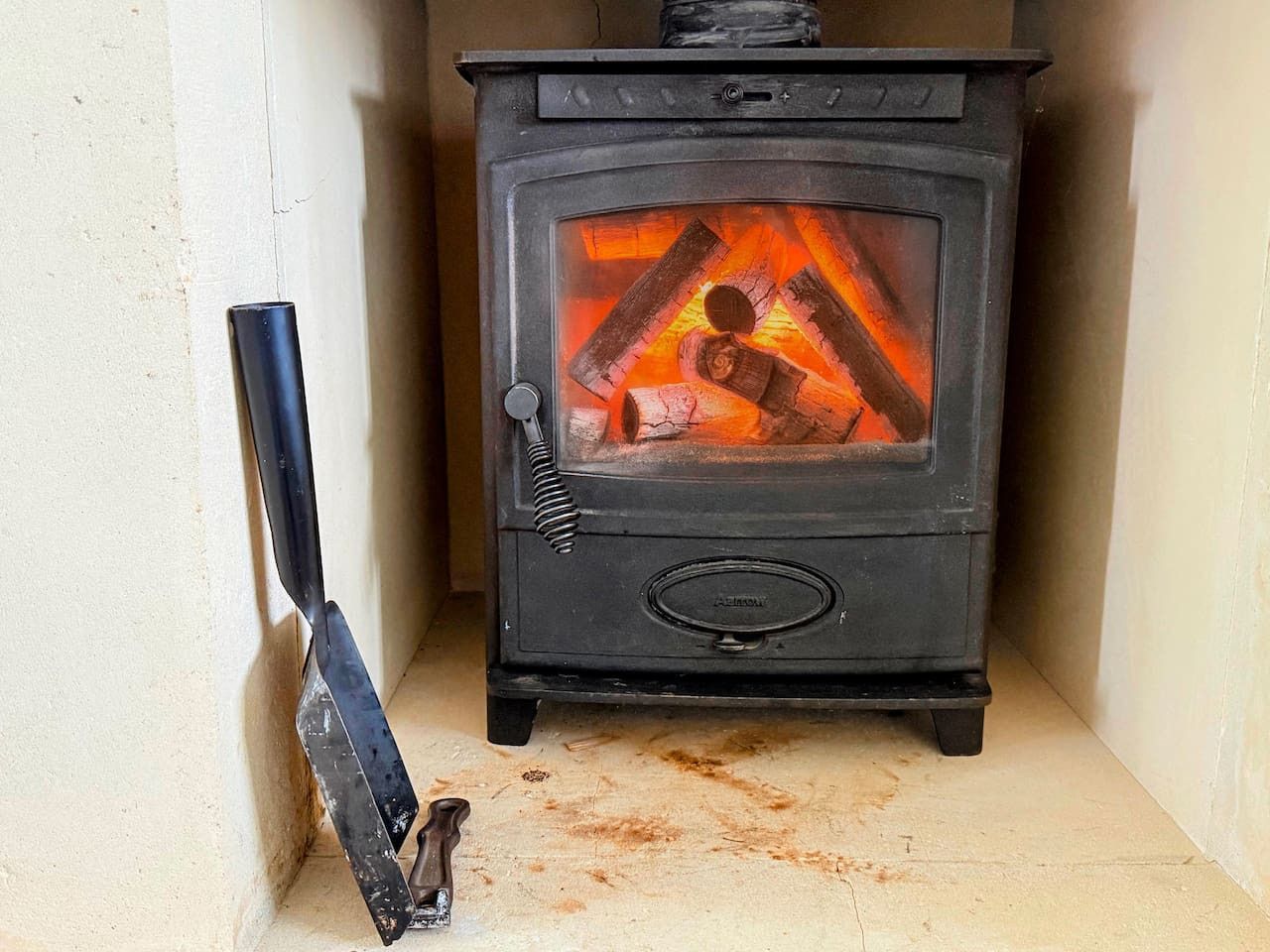 A black wood-burning stove with a glowing fire inside sits in a stone hearth, with a fireplace tool resting nearby.