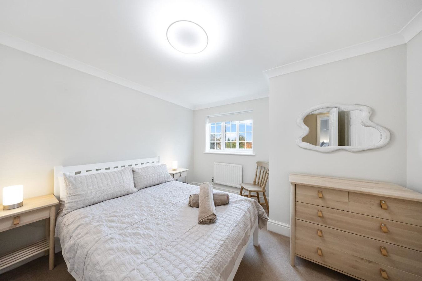 A bright, neutral-toned bedroom with a queen bed, light wood dresser, bedside table, and an organic-shaped wall mirror.