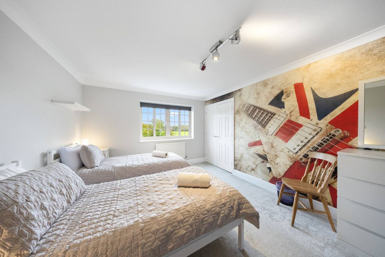 A bedroom with two beds and a Union Jack guitar wall mural, featuring a wooden chair and white dresser.