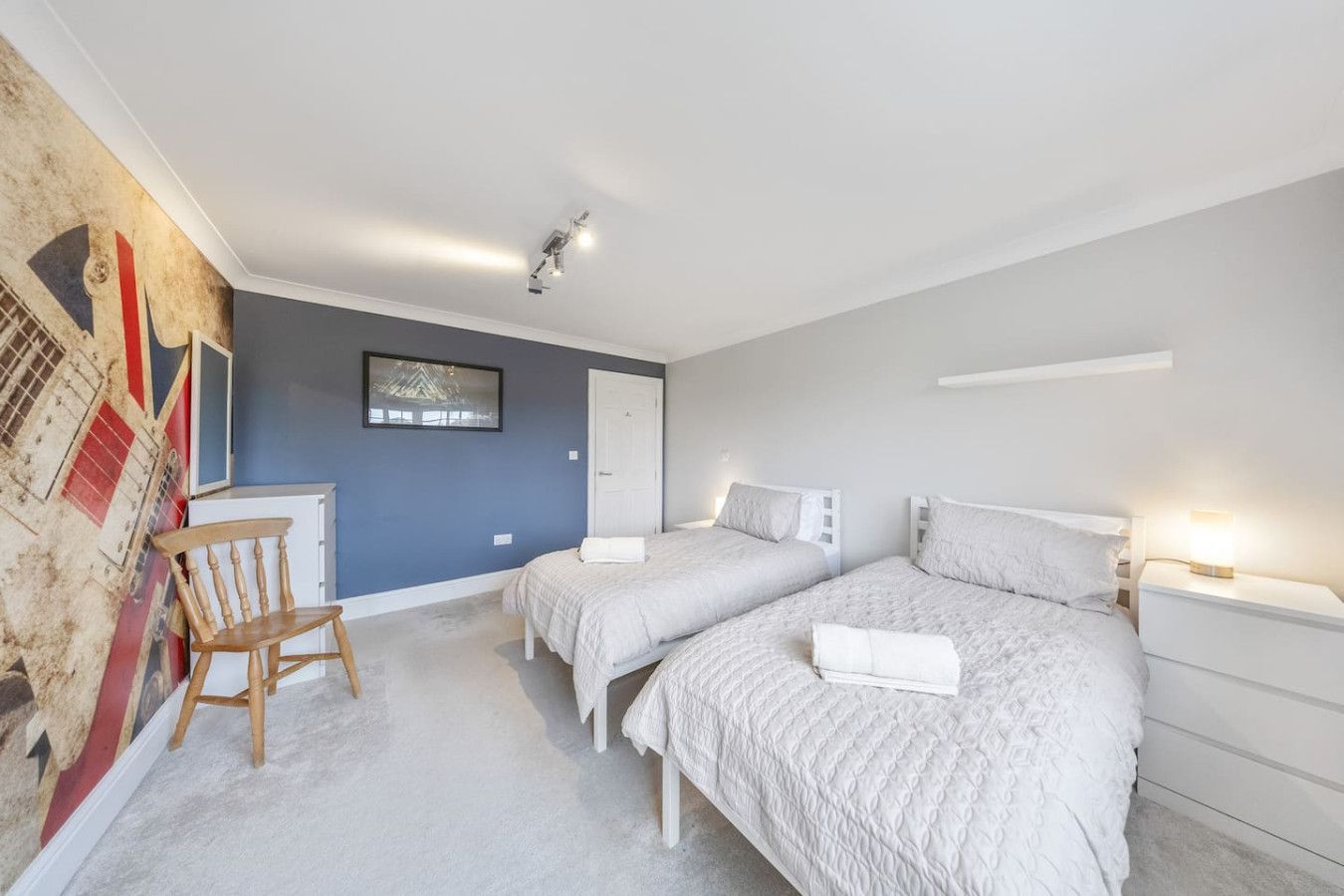 A twin bedroom featuring two beds with light-gray bedding, a wood chair, and a Union Jack feature wall against blue paint.