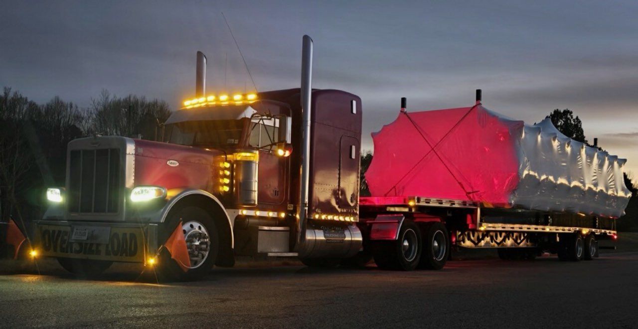 Purple Peterbilt semi-truck with a flatbed trailer loaded with concrete blocks parked outside a warehouse on a sunny day.
