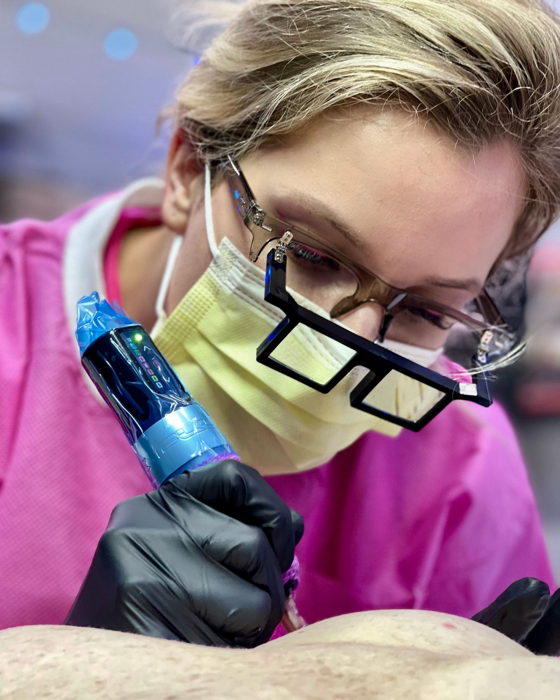 A woman wearing glasses and a mask is getting a tattoo on a person 's arm.
