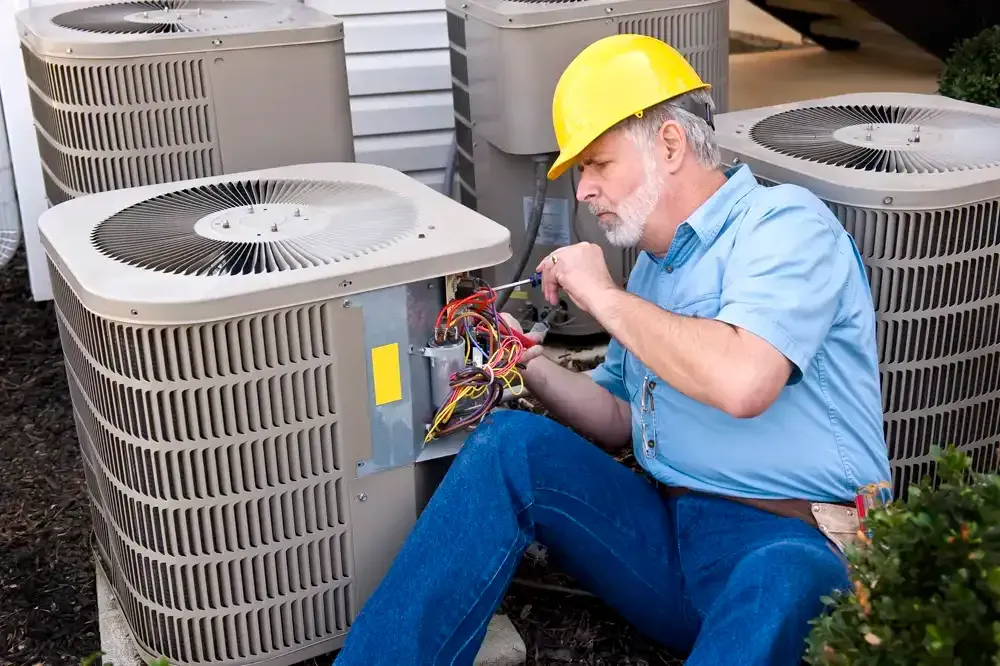 HVAC technician in a yellow hardhat repairs an air conditioning unit outdoors.