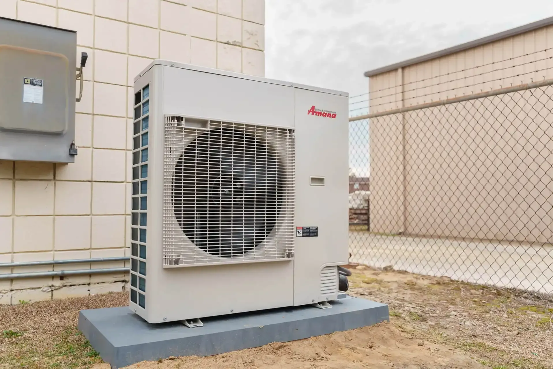 A beige Amada heat pump unit outside a building, with a chain link fence in the background.