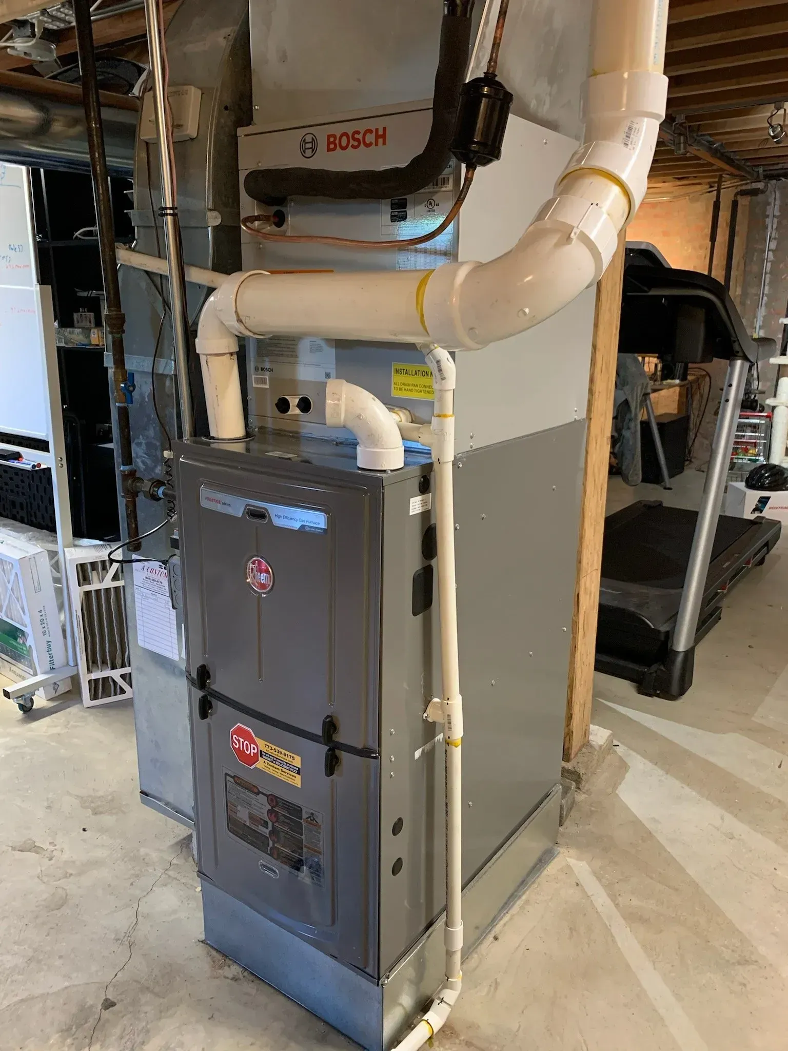 A Rheem furnace in a basement, with white pipes, grey metal, and a treadmill.