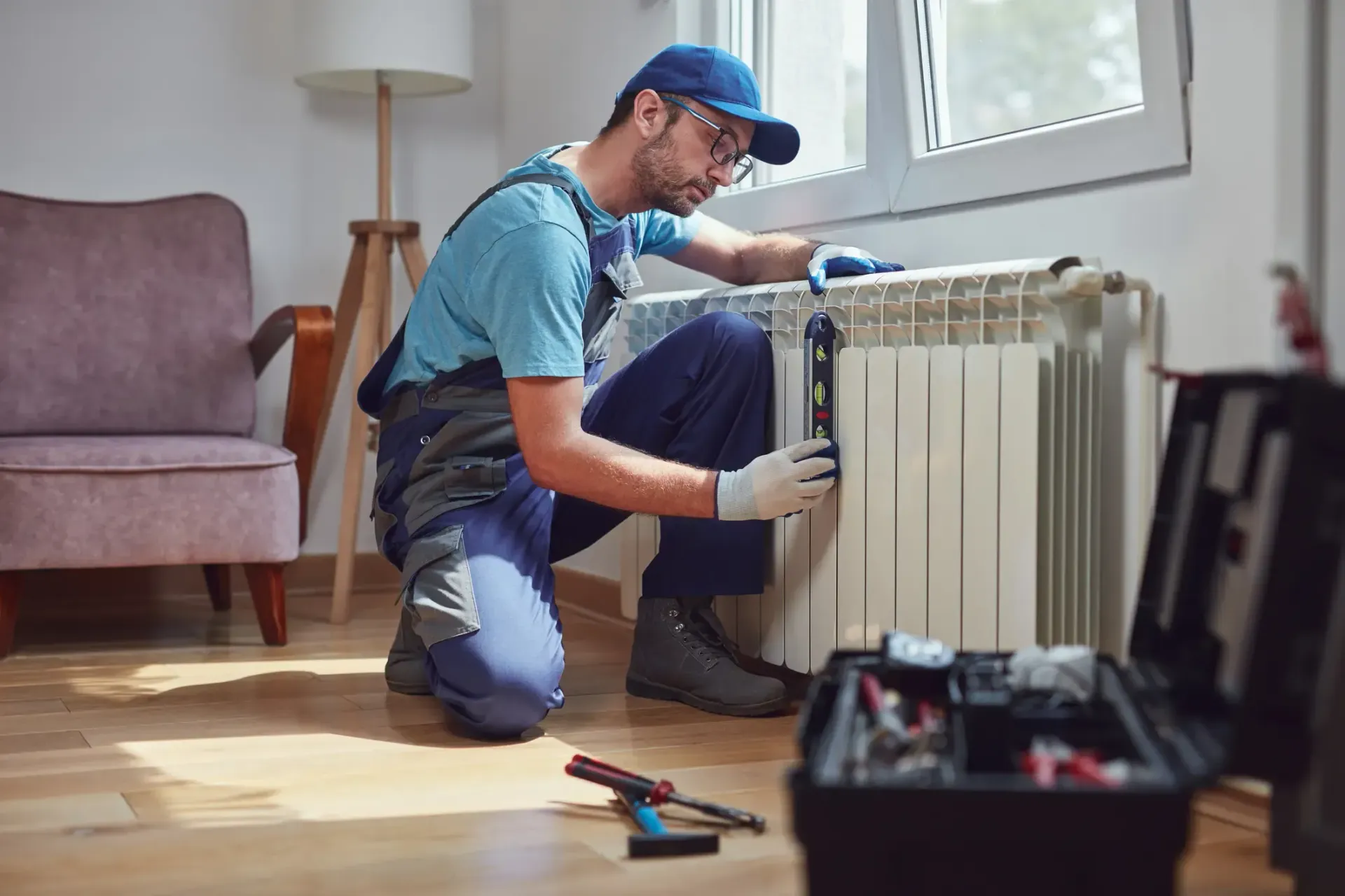A repairman kneels, fixing a radiator. He wears a cap, gloves, and overalls in a well-lit room.