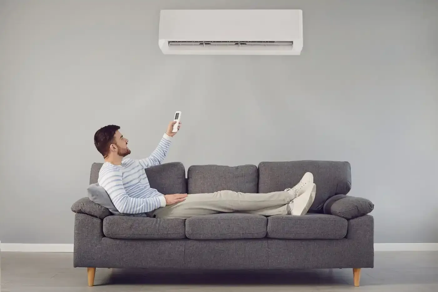 Man reclining on a gray sofa, using a remote control for the air conditioner on a light gray wall.