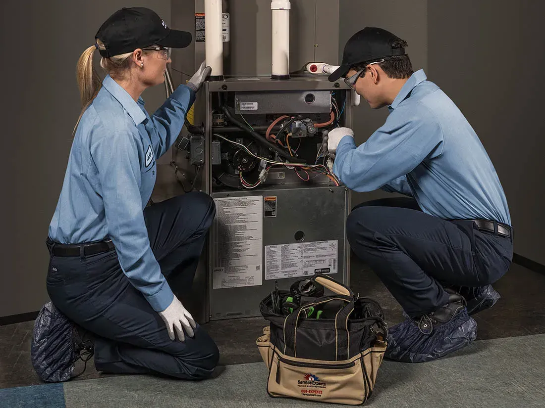 Two HVAC technicians in blue work uniforms servicing a furnace.