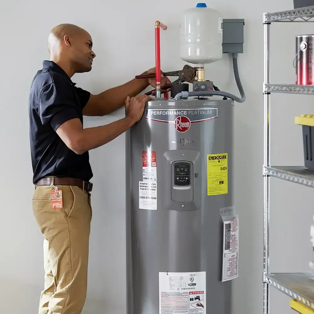A man in a black shirt adjusts a water heater in a garage, holding a copper pipe.