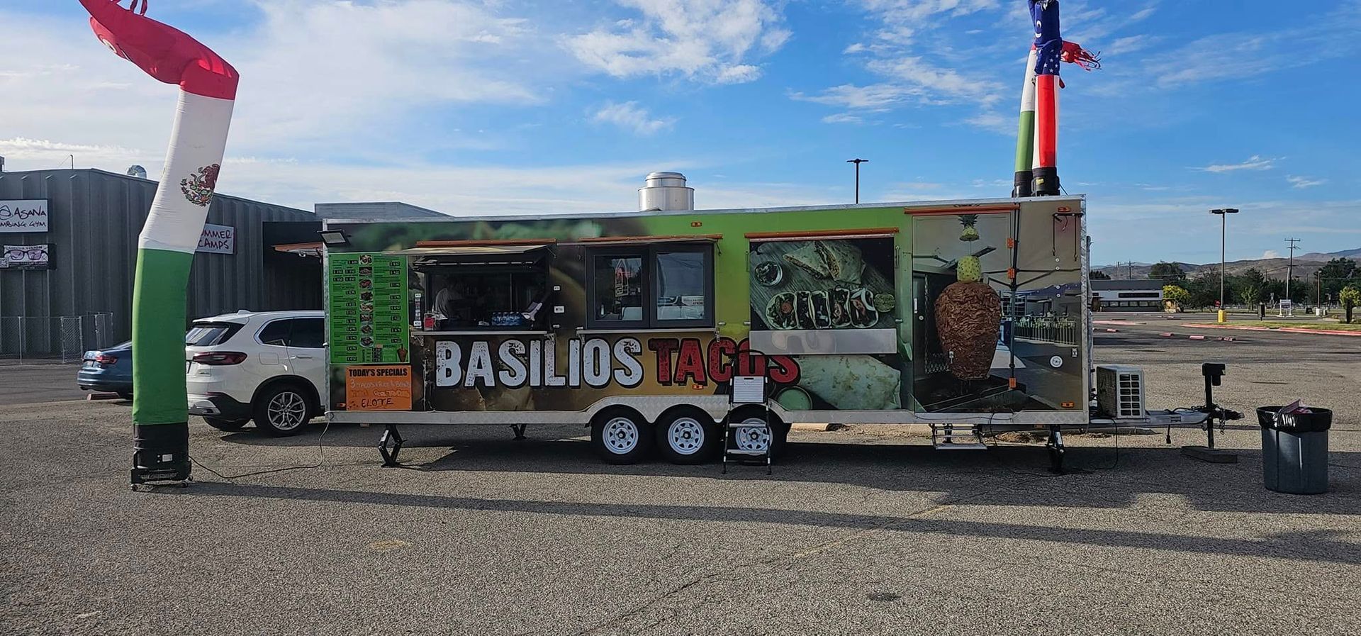 A food truck named Basilios Tacos with colorful air dancers in front, parked in a gravel lot under a cloudy sky.