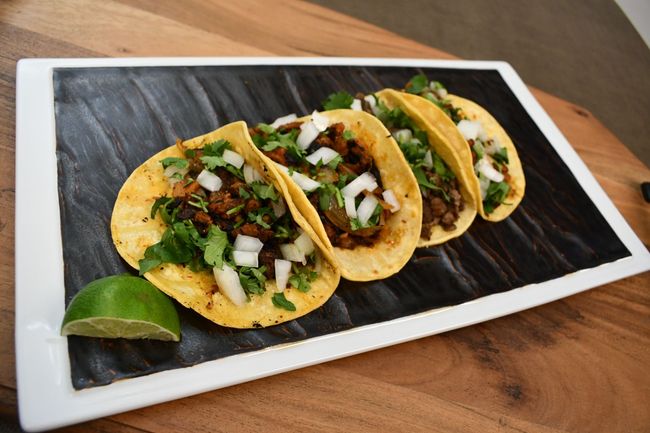 Three tacos filled with meat, cilantro, onions, avocado, and red sauce on a plate.