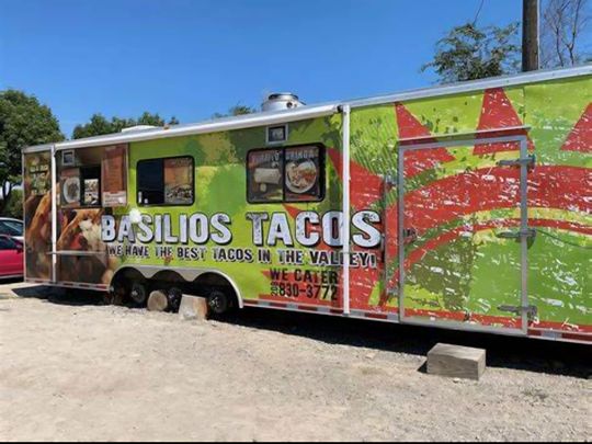 A food truck named Basilios Tacos with colorful air dancers in front, parked in a gravel lot under a cloudy sky.