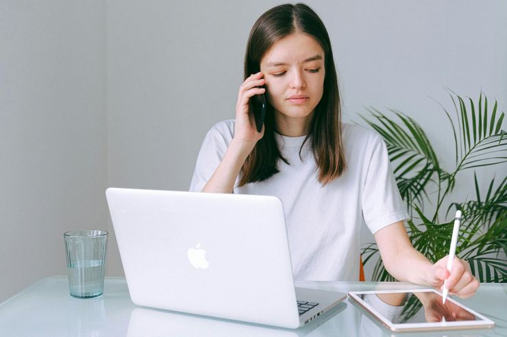 A woman is sitting at a desk with a laptop and a tablet while talking on a cell phone.