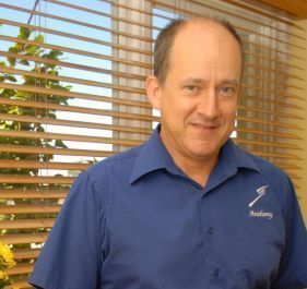 A man in a blue shirt is standing in front of a window with blinds.