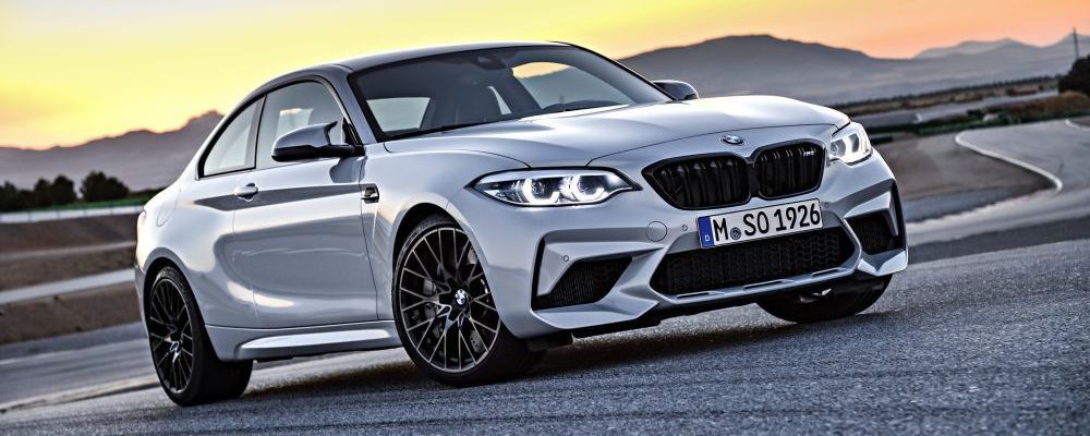 Een witte BMW M2 Competition staat bij een heuvelachtige weg met bergen op de achtergrond