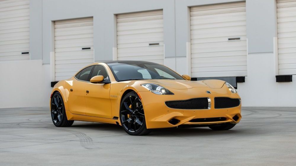 Een gele Fisker Karma staat op een terrein waar vrachtwagens laden en lossen